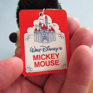 May include: A red tag with the words "Walt Disney's Mickey Mouse" and a castle illustration. The tag is attached to a black plush toy. The tag has a white border and is held in a hand.