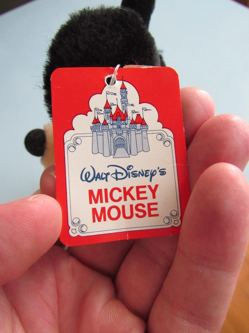 May include: A red tag with the words "Walt Disney's Mickey Mouse" and a castle illustration. The tag is attached to a black plush toy. The tag has a white border and is held in a hand.