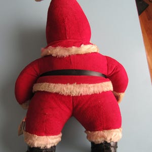 May include: A vintage Santa Claus doll, viewed from the back, wearing a red felt suit with white faux fur trim. The doll has a black belt and boots. The hat is tall and pointed, with a small yellow tassel.