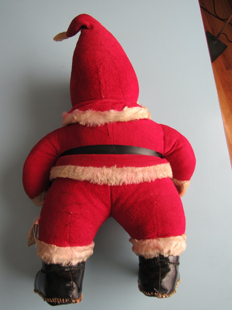 May include: A vintage Santa Claus doll, viewed from the back, wearing a red felt suit with white faux fur trim. The doll has a black belt and boots. The hat is tall and pointed, with a small yellow tassel.