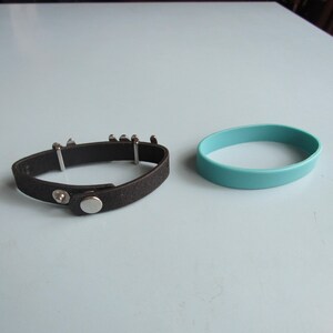 K&ouml;nnte beinhalten: Ein schwarzes Lederarmband mit silberfarbenen Metallakzenten und einem Druckknopfverschluss liegt neben einem hellblauen Silikonarmband. Die Armb&auml;nder liegen auf einer hellblauen Oberfl&auml;che.