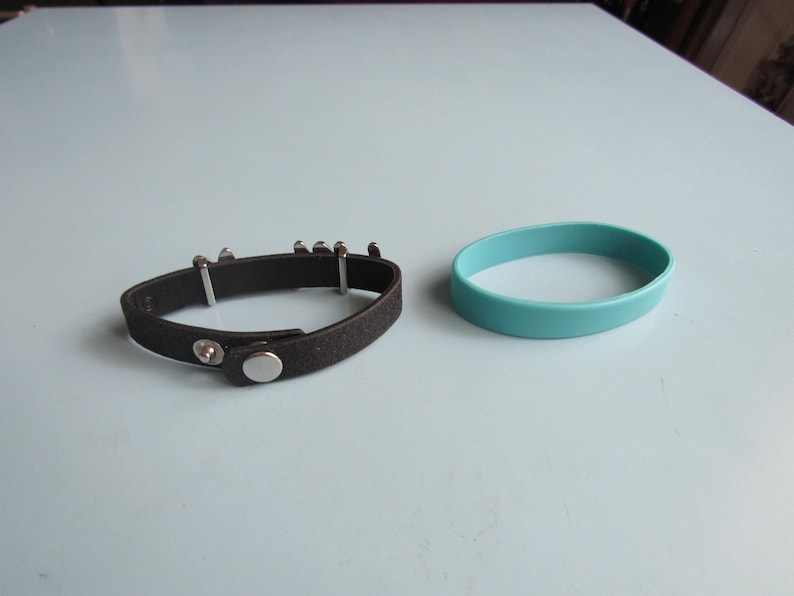 K&ouml;nnte beinhalten: Ein schwarzes Lederarmband mit silberfarbenen Metallakzenten und einem Druckknopfverschluss liegt neben einem hellblauen Silikonarmband. Die Armb&auml;nder liegen auf einer hellblauen Oberfl&auml;che.