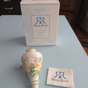 May include: A white porcelain decorative piece with a floral design and green beaded accents. The piece is next to a box and a small cloth, both with the "Rhyn-Rivét" logo and text. The box reads "A Collection Inspired by the Seasons."