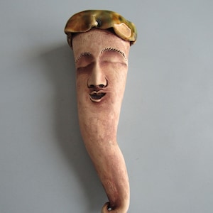 May include: A tall, slender ceramic sculpture featuring a human face with closed eyes and a small mouth. The surface has a matte, light brown finish. At the top, a glossy, rounded cap with mottled green and brown glaze sits like a hat.