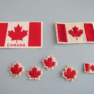 May include: A collection of Canadian flag and maple leaf patches. The flags are red and white, with a maple leaf in the center. One flag patch has the word "CANADA" printed on it. The maple leaf patches are red with white borders.