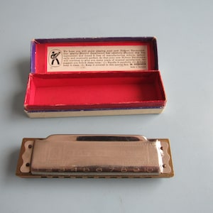 May include: A silver-colored Hohner harmonica with ten numbered holes and a metallic cover plate, accompanied by an open rectangular cardboard box. The box features a red interior and a white label with black text and a circular logo.