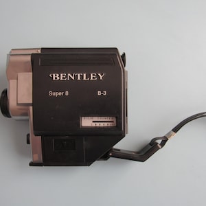 May include: A vintage Bentley Super 8 B-3 film camera. The camera is black and silver with the brand name and model details printed on the front. It has a film counter and a black strap attached to the side.