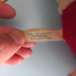 May include: A close-up of a vintage plush toy's tag. The tag reads "The Rushton Company, Atlanta, Ga." in a teal font. The toy is red and white, with a light blue background.