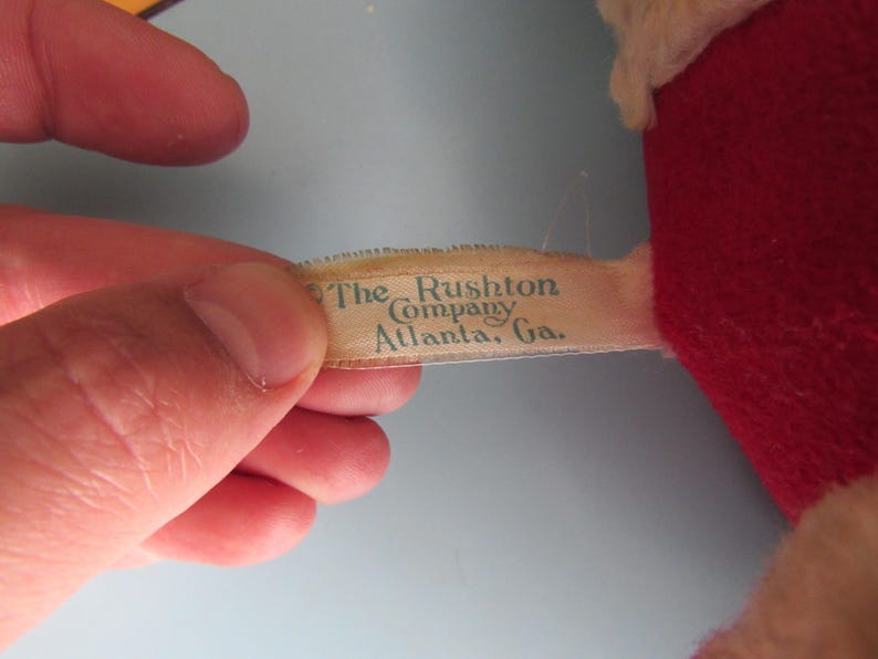 May include: A close-up of a vintage plush toy's tag. The tag reads "The Rushton Company, Atlanta, Ga." in a teal font. The toy is red and white, with a light blue background.