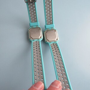 May include: Two digital watches with light blue straps and silver metal accents. The watch faces are square with a silver backing. The straps have a perforated design. The watches are displayed against a light blue background.
