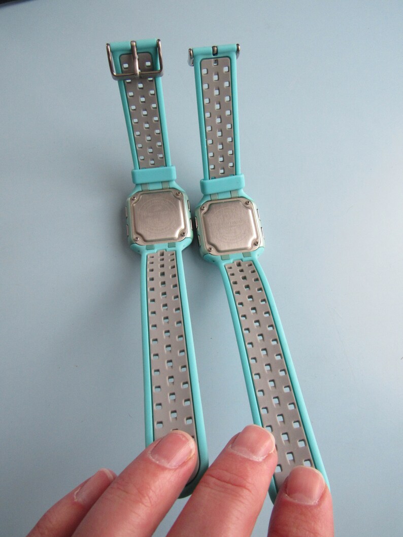 May include: Two digital watches with light blue straps and silver metal accents. The watch faces are square with a silver backing. The straps have a perforated design. The watches are displayed against a light blue background.