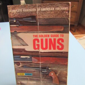 May include: A vintage book titled "The Golden Guide to Guns" with the text "Complete Handbook of American Firearms" at the top. The cover features illustrations of various firearms against a wood-grain background. The book is propped up on small, black feet.