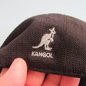 May include: A dark brown Kangol beret with a textured weave. The Kangol logo, featuring a kangaroo, is embroidered in light grey. The beret is a classic style, suitable for various occasions.
