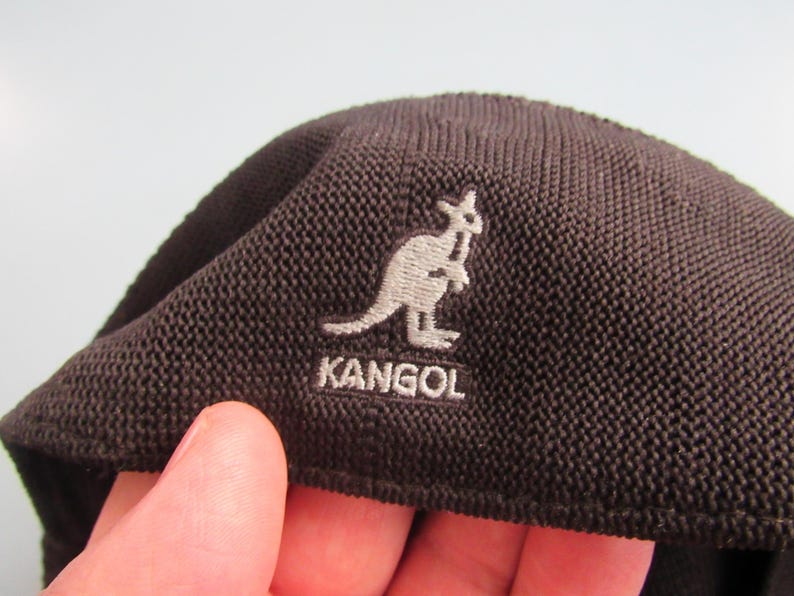 May include: A dark brown Kangol beret with a textured weave. The Kangol logo, featuring a kangaroo, is embroidered in light grey. The beret is a classic style, suitable for various occasions.