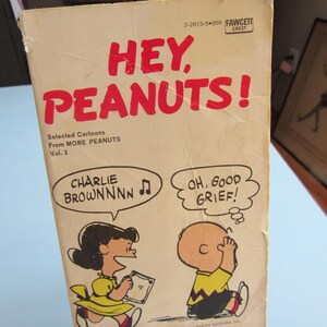 May include: A vintage paperback book cover featuring the Peanuts comic strip characters Charlie Brown and Lucy. The cover is yellow with red text that reads "Hey, Peanuts!" and "Selected Cartoons from MORE PEANUTS Vol. 2". The illustration shows Charlie Brown with his hands over his ears and Lucy singing with a music note above her head. The text in the speech bubbles reads "Charlie Brownnnn" and "Oh, Good Grief!"