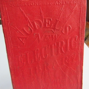May include: A red book with the title "Audels New Electric Library" embossed on the cover. The book has a decorative design with rays of light emanating from the center.