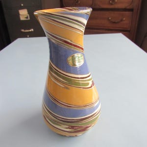 May include: A decorative glass vase with a swirling pattern of yellow, blue, green, and maroon stripes. The vase has a flared top and a rounded base. A gold sticker is affixed to the vase. The vase is approximately 8 inches tall.