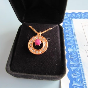 Puede incluir: Un collar de oro con un colgante circular con una gema rosa central rodeada de piedras transparentes más pequeñas. El collar se exhibe en una caja de terciopelo negro, con un certificado visible en el fondo.