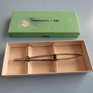 May include: An open, off-white cardboard box with a surgical instrument inside. The box is divided into three sections. A green box lid with the text "V. MUELLER & CO." is above the open box. The surgical instrument is silver.