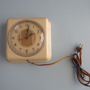 May include: Cream-colored, square-faced electric clock with a round clock face. The clock has a brown cord and plug. The clock face has black numbers and hands, with a brown center and a glass cover.