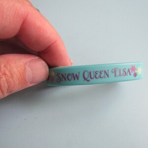 K&ouml;nnte beinhalten: Ein hellblaues Gummiarmband mit dem Text "SNOW QUEEN ELSA" in Lila. Das Armband hat dekorative Blumen- und Sternakzente. Es ist ein Kinderaccessoire.