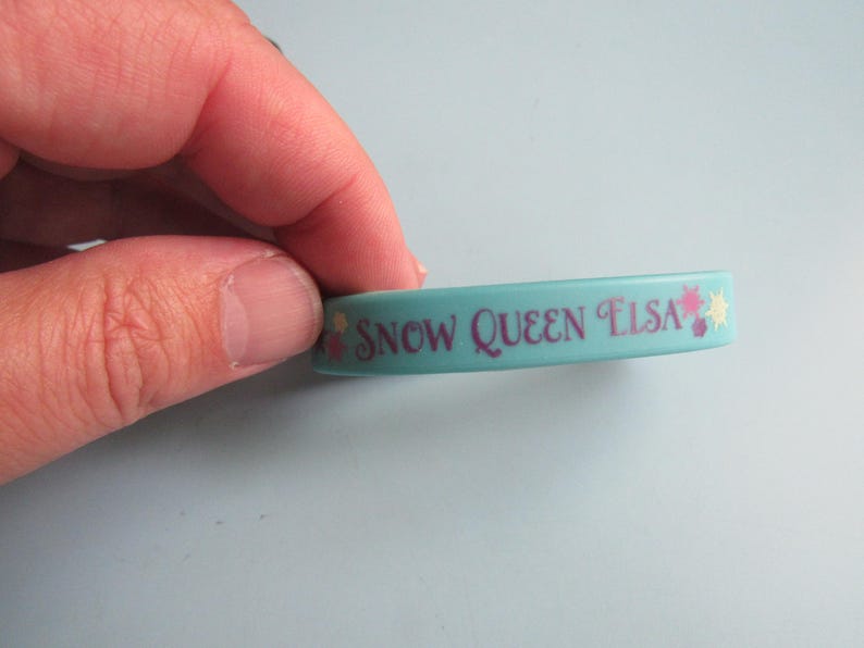 K&ouml;nnte beinhalten: Ein hellblaues Gummiarmband mit dem Text "SNOW QUEEN ELSA" in Lila. Das Armband hat dekorative Blumen- und Sternakzente. Es ist ein Kinderaccessoire.