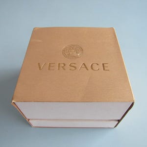 May include: A square, gold-colored box with a textured surface and a white base. The top of the box features the embossed Versace logo and the word VERSACE in capital letters centered on the lid. The box is closed and viewed from above.