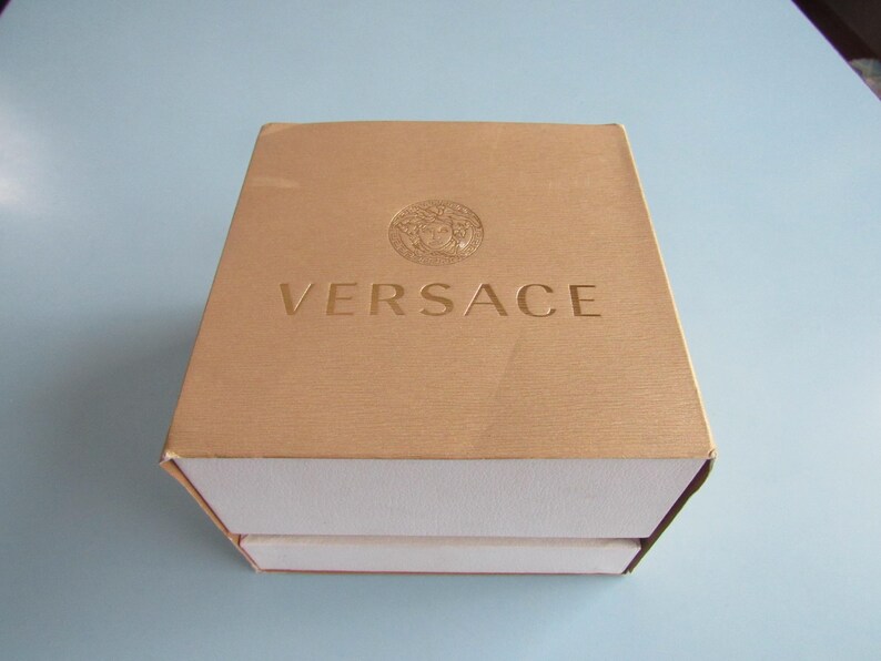 May include: A square, gold-colored box with a textured surface and a white base. The top of the box features the embossed Versace logo and the word VERSACE in capital letters centered on the lid. The box is closed and viewed from above.