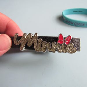 K&ouml;nnte beinhalten: Ein schwarzes Glitzerarmband mit dem Namen "Minnie" in silbernen Buchstaben mit Glitzerakzenten. Eine rote Schleife ist auf der rechten Seite des Namens angebracht. Ein hellblaues Gummiband mit Text befindet sich im Hintergrund.