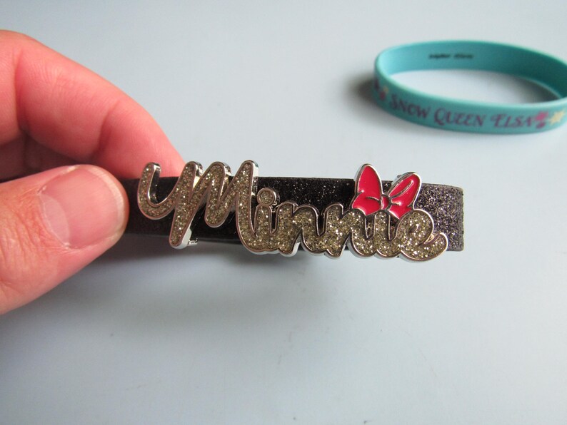 K&ouml;nnte beinhalten: Ein schwarzes Glitzerarmband mit dem Namen "Minnie" in silbernen Buchstaben mit Glitzerakzenten. Eine rote Schleife ist auf der rechten Seite des Namens angebracht. Ein hellblaues Gummiband mit Text befindet sich im Hintergrund.