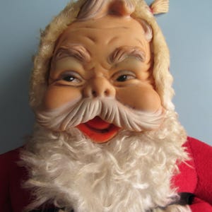 May include: A vintage Santa Claus doll with a detailed, expressive face. The doll features a tan face, white beard, and a red suit. The Santa Claus doll has a fluffy, light-colored beard and a red mouth. The doll is a classic Christmas decoration.