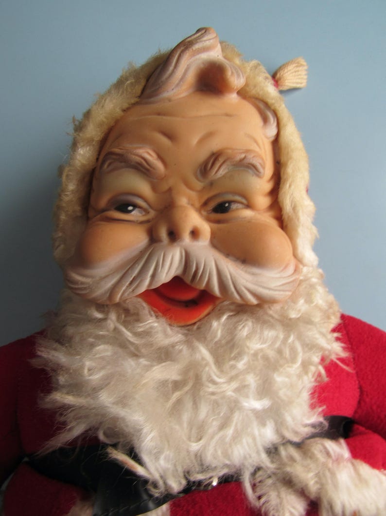May include: A vintage Santa Claus doll with a detailed, expressive face. The doll features a tan face, white beard, and a red suit. The Santa Claus doll has a fluffy, light-colored beard and a red mouth. The doll is a classic Christmas decoration.