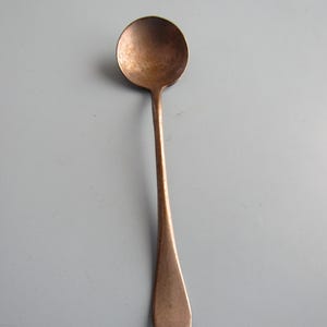 May include: A vintage copper-colored spoon with a round bowl and a long, slender handle. The spoon has a slightly tarnished appearance, suggesting age and use. The handle tapers to a rounded end. The spoon is likely for serving or eating.