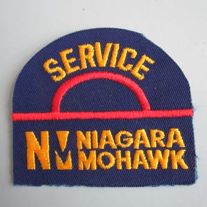 May include: Embroidered patch with a navy blue background. The patch features the word "SERVICE" in yellow arched across the top, with a red semi-circle and line beneath it. Below, the text "NIAGARA MOHAWK" is in yellow.