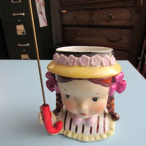 May include: A vintage ceramic figurine of a girl with brown braids, wearing a yellow hat with pink flowers and holding a red umbrella. The figurine has a painted face and is wearing a pink bow. The base is striped.