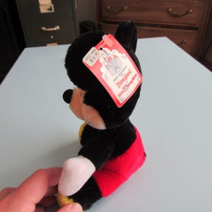 May include: A plush Mickey Mouse toy, primarily black with red shorts, white gloves, and yellow shoes. The toy has a red tag that reads "Made Exclusively for Disneyland". The toy is sitting on a light blue surface.