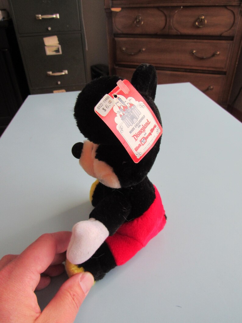 May include: A plush Mickey Mouse toy, primarily black with red shorts, white gloves, and yellow shoes. The toy has a red tag that reads "Made Exclusively for Disneyland". The toy is sitting on a light blue surface.