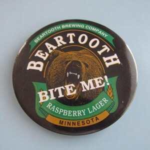 May include: A round, dark brown beer coaster with the text "BEARTOOTH BREWING COMPANY" in white. The center features a bear's head with the words "BITE ME!" and "RASPBERRY LAGER MINNESOTA".