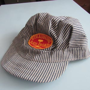 May include: A blue and white hickory striped engineer cap featuring a prominent oval patch on the front. The patch has a red background with yellow text that reads Strasburg Rail Road and includes a yellow graphic of a railroad spike.