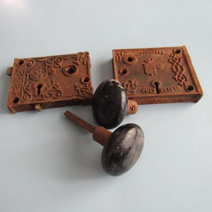 May include: Antique door hardware set, featuring two rusty, rectangular lock plates with keyholes and decorative patterns. Two black oval doorknobs with brown stems are also included. The hardware is likely made of cast iron.