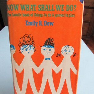 May include: A hardcover book with a bright orange cover titled Now What Shall We Do? The family book of things to do & games to play by Emily R. Dow. The cover features a line drawing of four stylized white figures holding hands in blue ink.