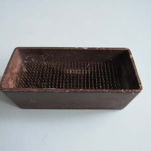 May include: A rectangular brown floral frog featuring a heavy base with a series of upright metal pins arranged in rows. The container has a matte finish with visible signs of wear along the top edges and a deep interior holding the needle points.