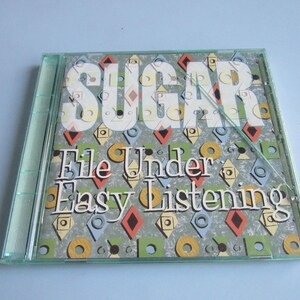 May include: A clear plastic CD jewel case containing the album Sugar, File Under Easy Listening. The cover art features a gray background with a repeating pattern of colorful geometric shapes including red diamonds, yellow circles, and blue squares.