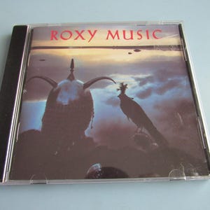 May include: A compact disc case featuring the band name "Roxy Music" in red lettering. The album art depicts a dark, silhouetted figure with a horned helmet and a bird against a cloudy sky.