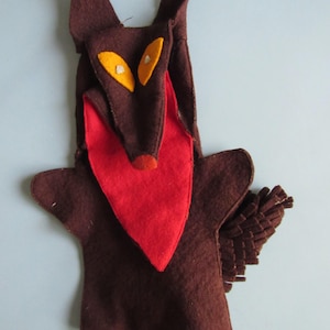 May include: A brown felt hand puppet shaped like a wolf with pointed ears, yellow eyes, a red chest patch, and a bushy tail made of layered felt strips. The puppet features a long snout with a small red nose and a wide, open mouth design.