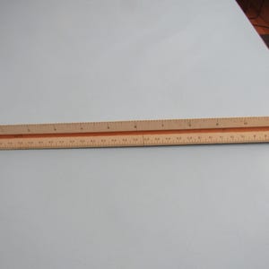 May include: A wooden architectural scale ruler with markings in inches. The ruler is light brown with black and red markings. It is lying on a light blue surface.