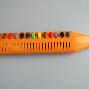 May include: An orange melodica with a series of colored keys. The instrument has a rectangular body with vertical slits and a mouthpiece at one end. The keys are brown, red, purple, blue, green, yellow, brown, purple, black, and blue.