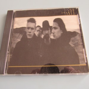 May include: A U2 CD case featuring a sepia-toned image of the band members. The album title "The Joshua Tree" is printed in white at the top. The case is in a clear plastic jewel case with a brown spine.