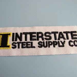 May include: A rectangular white patch with the text "INTERSTATE STEEL SUPPLY CO." embroidered in black. The letter "I" is black with a yellow outline. The patch is likely for a work uniform or apparel.