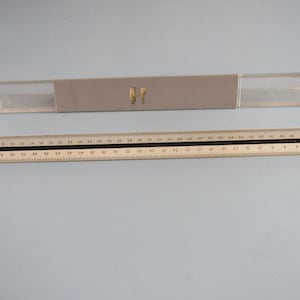 May include: A white ruler with black markings, measuring up to 75 cm, with a gray plastic case with the letters "RF" in gold.